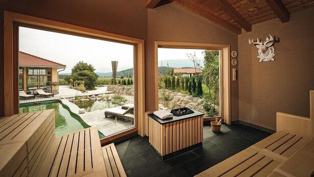Um espaço de spa com um banco de madeira, uma pequena mesa com uma toalha e um balde, e vista para uma piscina e jardim através de amplas janelas.