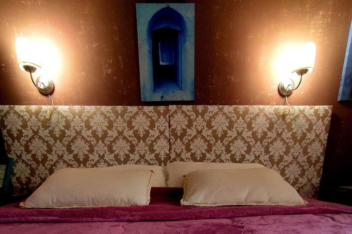 Une chambre d’hôtel avec une tête de lit à motifs, deux lampes de chevet, un lit recouvert d’une couverture rouge et un coussin décoratif.