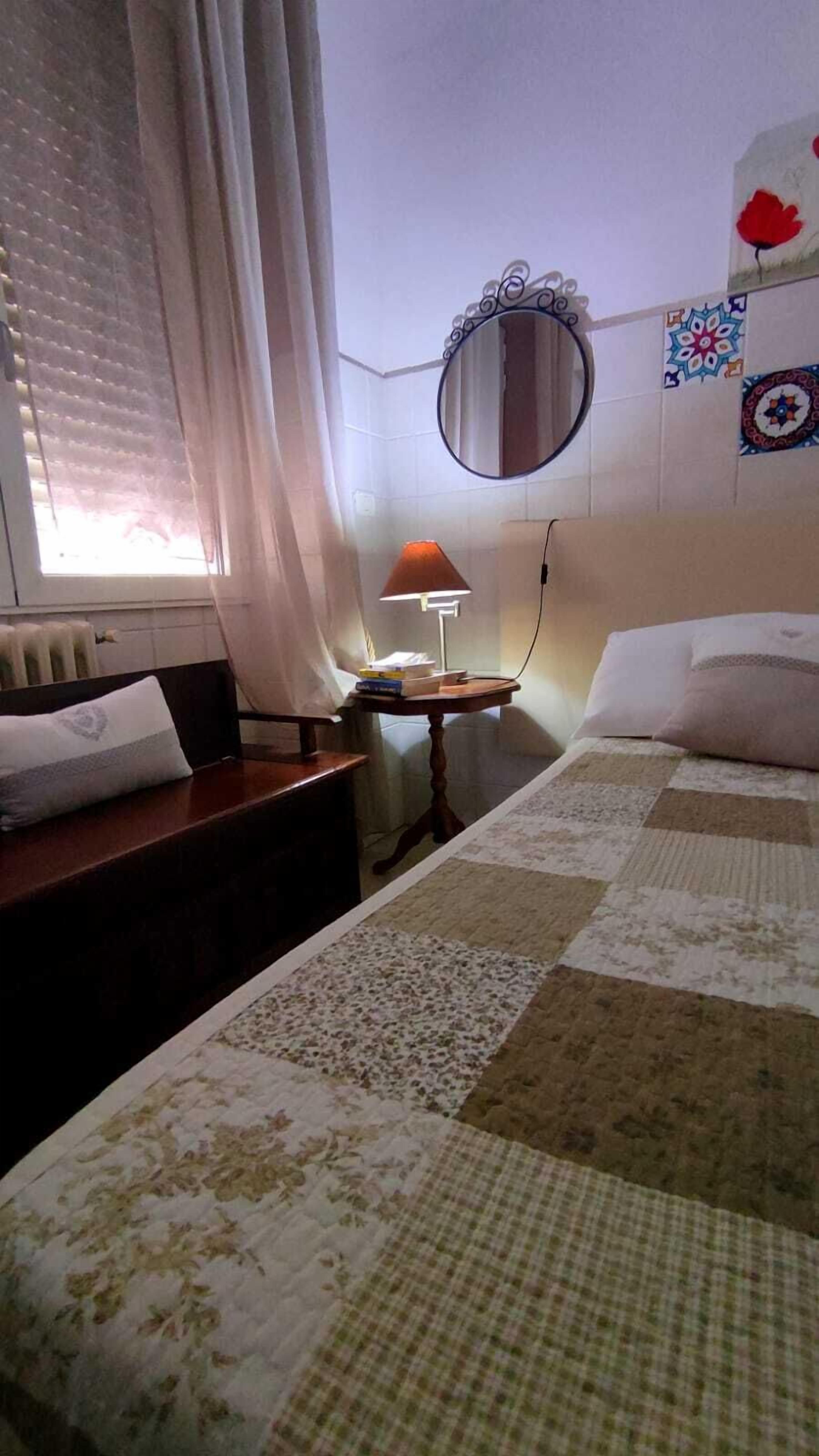 A hotel room with a bed, a bedside table with a lamp, a mirror, and a window with curtains.