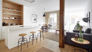 A modern kitchen with white cabinetry, wooden shelves, and stools. There is a dining area with a table and chairs, and a living room with a sofa and a small side table. - Miramon by FeelFree Rentals (San Sebastián)
