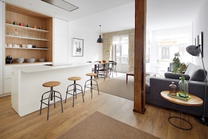 Una cocina moderna con armarios blancos, estantes de madera y taburetes. Hay un comedor con una mesa y sillas, y una sala de estar con un sofá y una pequeña mesita.