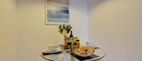 Een klein eethoekje met een glazen tafel, twee krukken, een schaal eten, een fles wijn en een vaas bloemen.