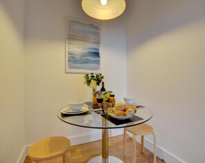 A small dining area with a glass table, two stools, a bowl of food, a bottle of wine, and a vase of flowers.