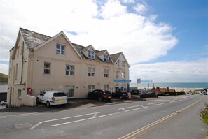 A two-story building with a flat roof and a blue awning. There are cars parked in front, and a beach is visible in the background. - 1 Bed in Woolacombe (oc-seablu) (Woolacombe)