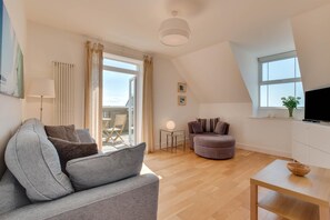 En moderne stue med en grå sofa, en rund skammel, et lille bord og et fjernsyn på et hvidt skab. Der er en altan med en stol og et bord, samt et vindue med en plante i vindueskarmen.