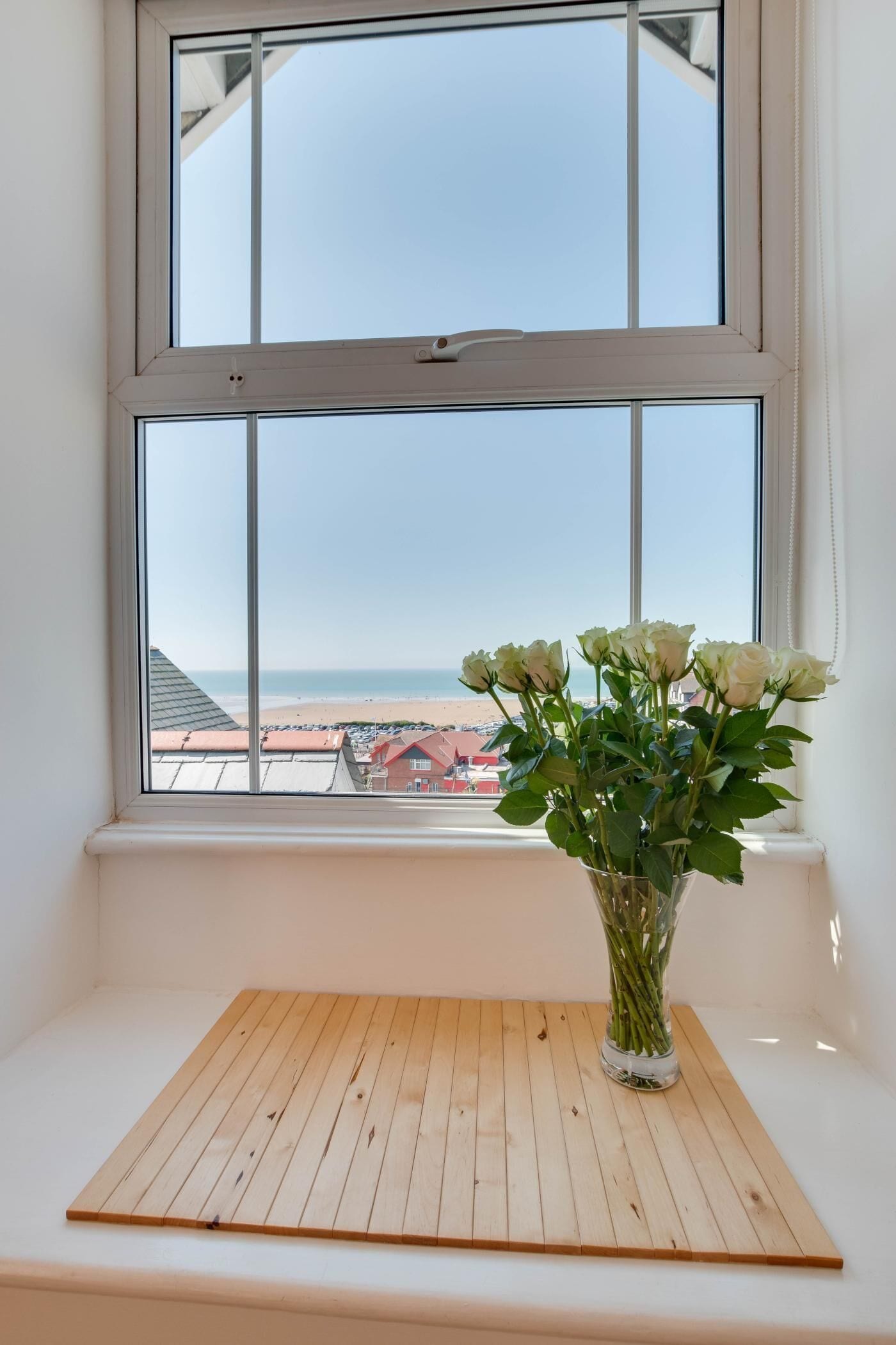 Una finestra con vista sulla spiaggia e un vaso con fiori bianchi su un ripiano di legno.