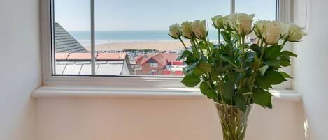 Una finestra con vista sulla spiaggia e un vaso con fiori bianchi su un ripiano di legno.