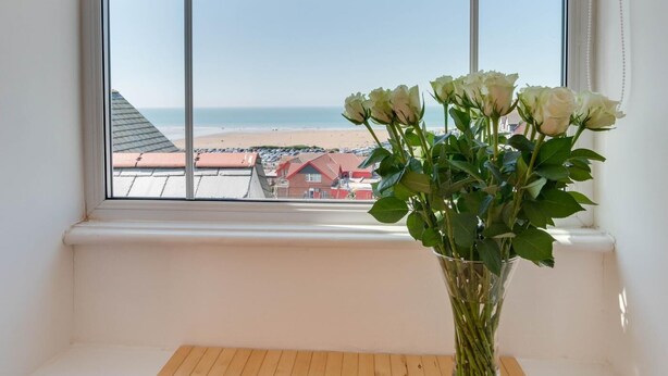 A window with a view of a beach and a vase with white flowers on a wooden ledge. - 1 Bed in Woolacombe (oc-seablu) (Woolacombe)