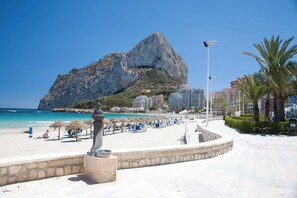 A beach with a rocky mountain in the background, a walkway with umbrellas, and buildings nearby.