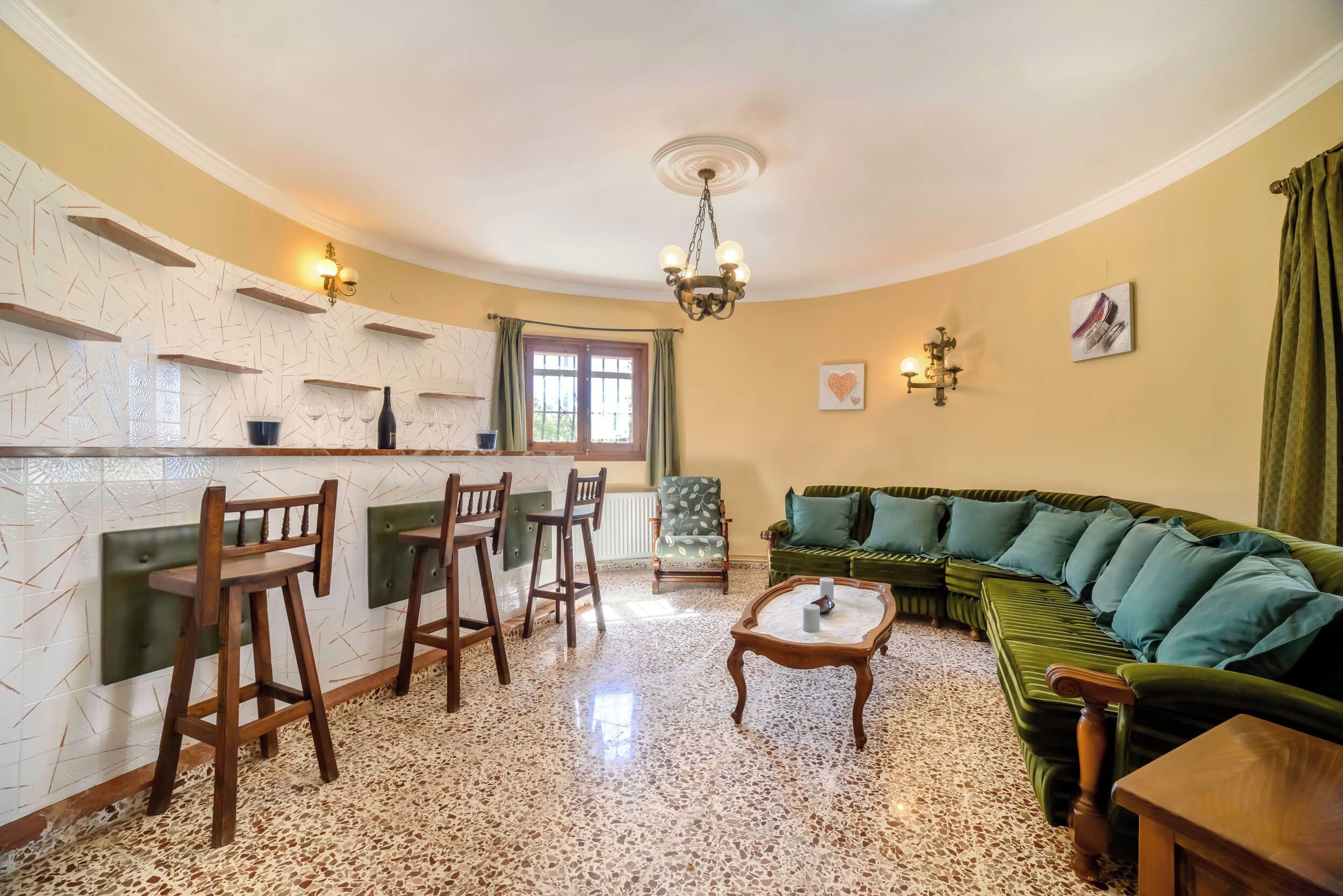Un salon avec un canapé vert, des chaises en bois, une table basse et un coin cuisine doté d’un comptoir-bar et d’étagères.