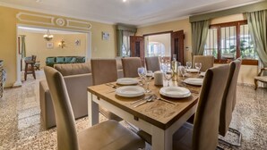 A dining area with a wooden table set for four, surrounded by chairs. There is a sofa and a dining table in the background. - Villa Puchero - PlusHolidays (Calpe)