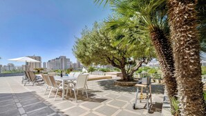 Un espace extérieur aménagé avec des tables et des chaises, un palmier, et une vue sur le paysage urbain en arrière-plan.
