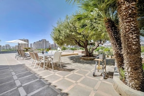 An outdoor seating area with tables and chairs, a palm tree, and a view of the cityscape in the background.