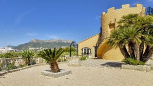 A building with a yellow facade, a stone tower, and a paved courtyard with palm trees and planters. - Villa Puchero - PlusHolidays (Calpe)