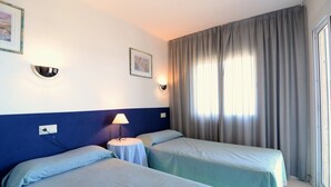 Habitación de hotel con dos camas individuales, una mesita con una lámpara, una luz en la pared y un ventanal con cortinas.