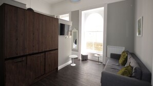 A modern living room with a dark wood wardrobe, a grey sofa with cushions, a small white side table, and a large window with a view of a stone wall. - RoomApart studio sleeps 2-4 on the ground floor (Plymouth)