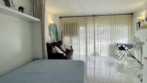 Een kamer met een bed, een slaapbank, een tafel met een ventilator en een raam met gordijnen.