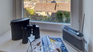 Une fenêtre donnant sur une ville côtière, une paire de jumelles, une radio et un magazine sur l’observation des oiseaux, le tout posé sur un rebord de fenêtre.