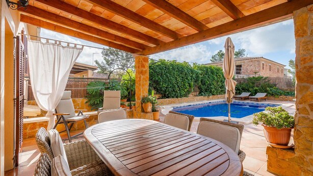 An outdoor patio with a wooden table and chairs, a pool, and a stone building in the background.