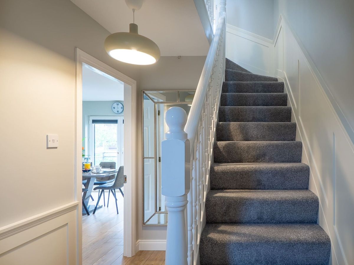 A staircase with carpeted steps and white balusters, leading to a room with a dining area and a clock on the wall.