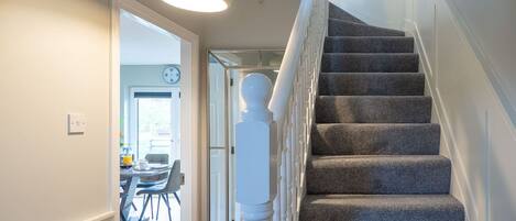 A staircase with carpeted steps and white balusters, leading to a room with a dining area and a clock on the wall.