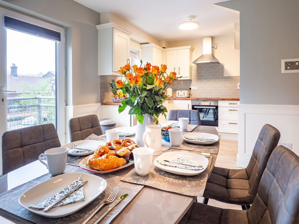 A dining area with a table set for breakfast, including pastries and coffee, with a view of the kitchen in the background.