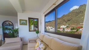 Una sala de estar con sofá y sillón, una mesita y una ventana que ofrece vistas a un pueblo en la ladera.