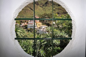 Vista a través de una ventana circular con diseño de cuadrícula, que muestra una ladera con edificios y abundante vegetación.