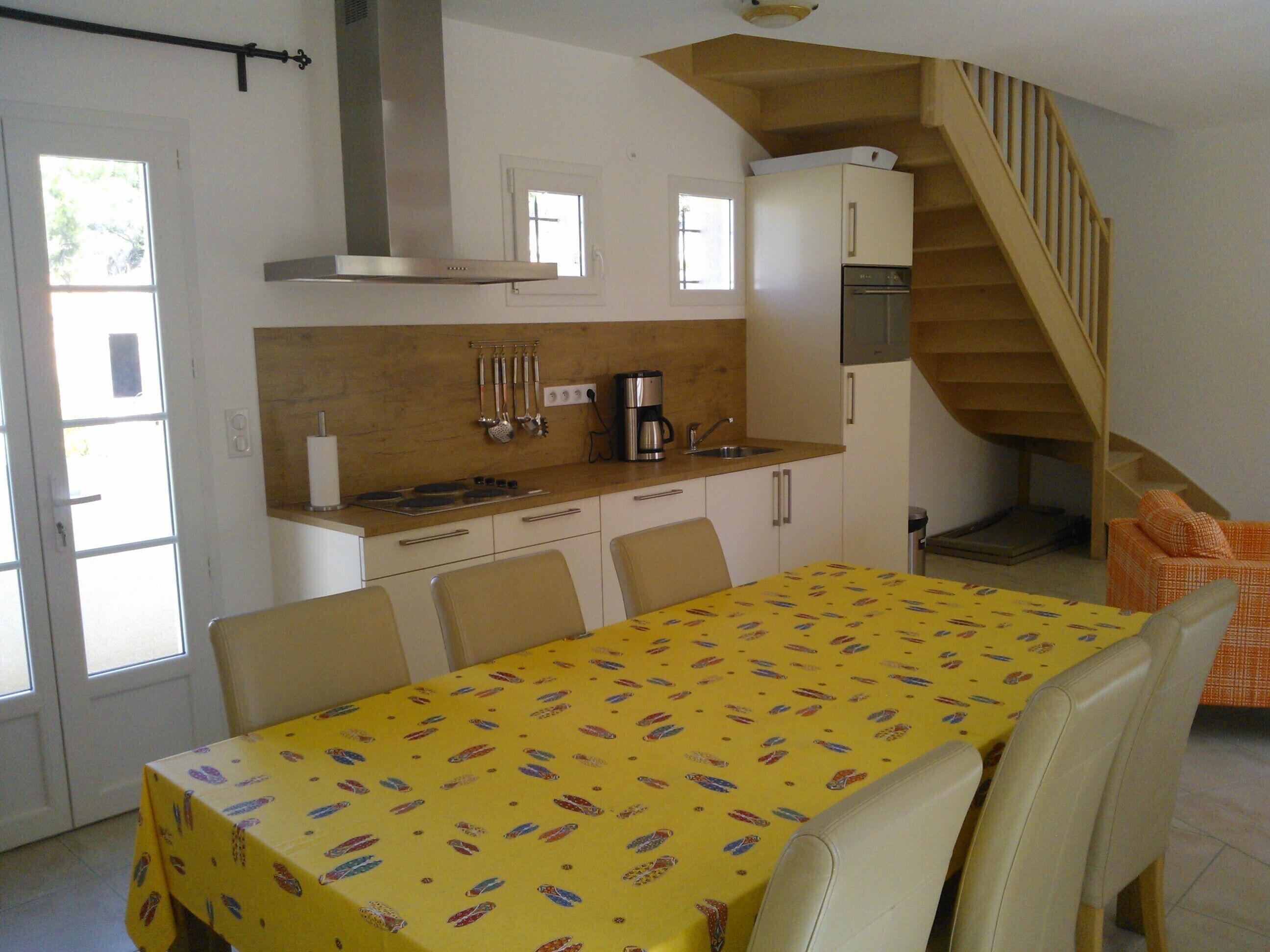 Une cuisine compacte avec un coin repas, agrémentée d’une nappe jaune, de meubles blancs et d’un escalier en bois.