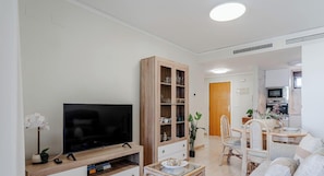 A modern living room with a flat-screen TV, wooden cabinet, dining area with chairs, and a kitchen in the background.