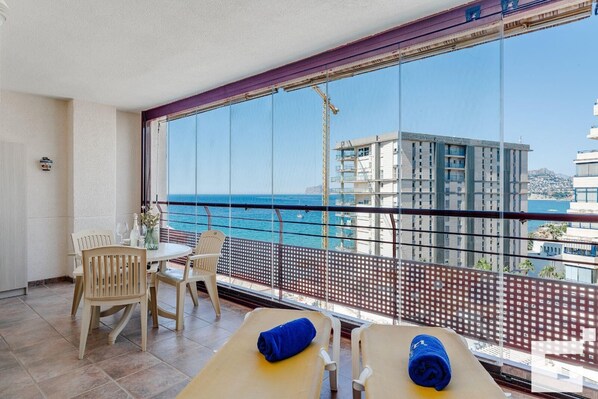 A balcony with a view of the sea and buildings, a table with chairs, and a massage table with towels.