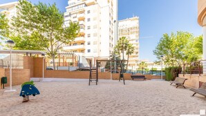 A sandy playground with swings, a slide, and a climbing structure, surrounded by buildings and trees. - Nautilus 39B - Grupo Turis - Calpe, Costa Blanca (Calpe)