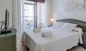 A hotel room with a bed, a TV on a stand, a chair, a table, and a balcony with a view.