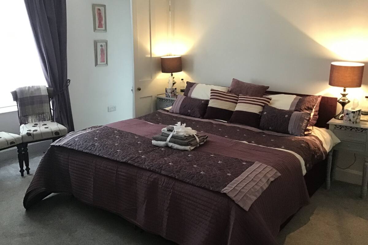 A neatly made bed with a brown quilt, two bedside lamps, a chair with a blanket, and a door.