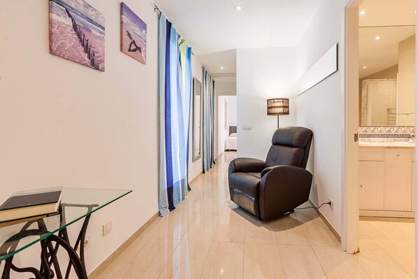 A modern hotel room with a massage chair, a glass table with a book, and a view of the hallway.