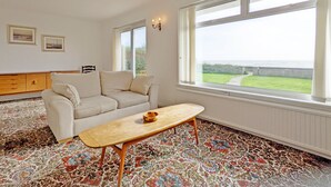 Una habitación con un sofá color beige, una mesa de centro de madera y una alfombra estampada. Hay dos ventanas con vistas al exterior.