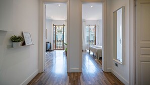 Un pasillo con piso de madera, paredes blancas y una puerta que da a una habitación con cama y ventana.