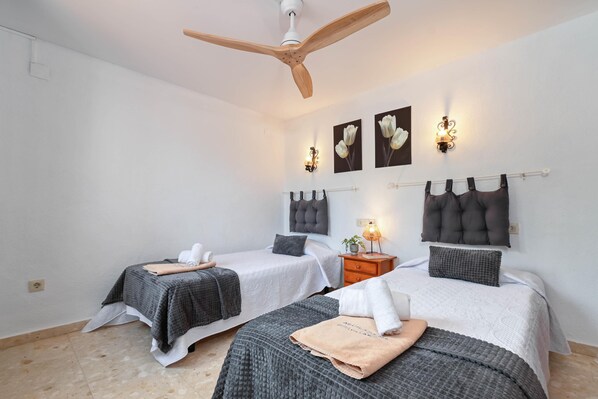 A hotel room with two beds, a ceiling fan, wall-mounted light fixtures, and a small bedside table with a lamp.