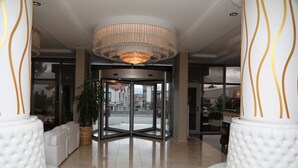 En foyer med en drejeomdrejningsdør, hvide polstrede siddepladser, en stor lysekrone og en potteplante.