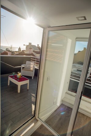 A balcony with a white chair and a small table, a glass door, and a view of buildings in the background.