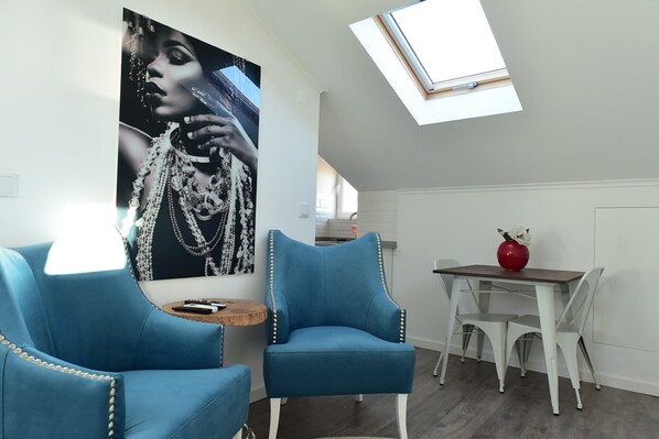 A modern living room with a skylight, a blue armchair, a dining table with white chairs, and a large poster on the wall.