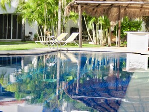 A swimming pool with clear water reflecting palm trees and a thatched-roof structure. - Playacar -Executive Comfort Room With Swimming Pool Air Conditioning and Parking (Playa del Carmen)