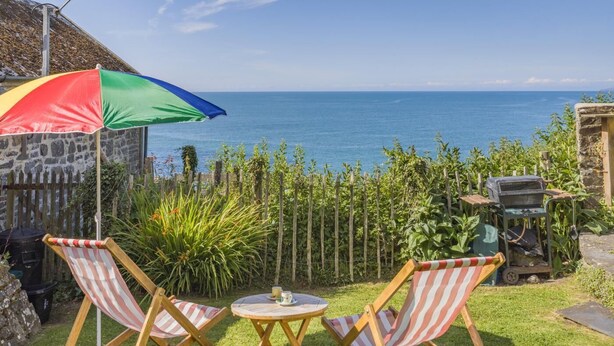 A patio with two striped lounge chairs, a small round table, and a colorful umbrella. - 2 Bed in Bideford (oc-willi) (Buck's Mills)