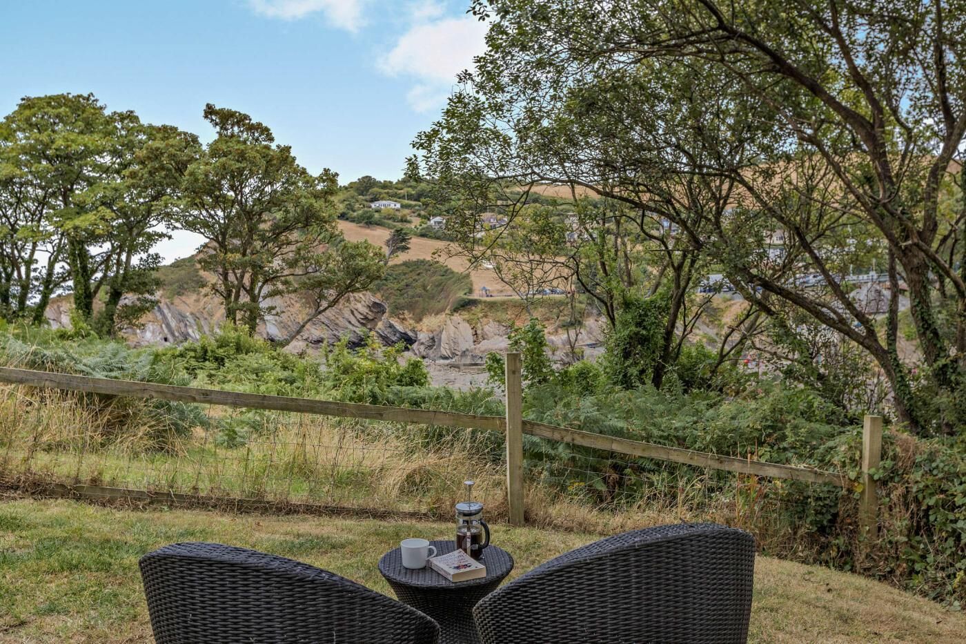 Ein Korbstuhl mit einem Buch und einer Tasse, mit Blick auf eine natürliche Landschaft mit Bäumen und felsigen Hügeln.
