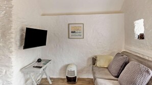 A room with a television mounted on the wall, a small table with a remote control, a heater, a sofa with cushions, and a framed picture on the wall.