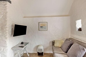 A room with a television mounted on the wall, a small table with a remote control, a heater, a sofa with cushions, and a framed picture on the wall.