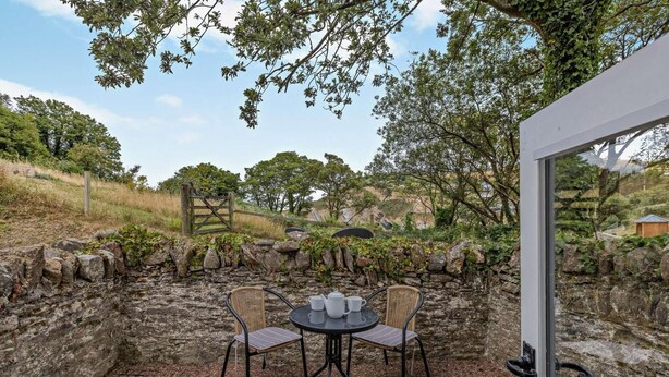An outdoor patio with stone walls, two wicker chairs, a table with cups, and a view of a natural landscape.