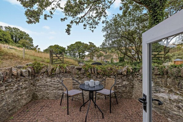 An outdoor patio with stone walls, two wicker chairs, a table with cups, and a view of a natural landscape.