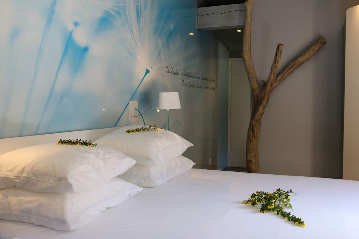 Una cama bien hecha con sábanas blancas y flores decorativas, una pared con un diseño abstracto azul y una rama de madera a la derecha.
