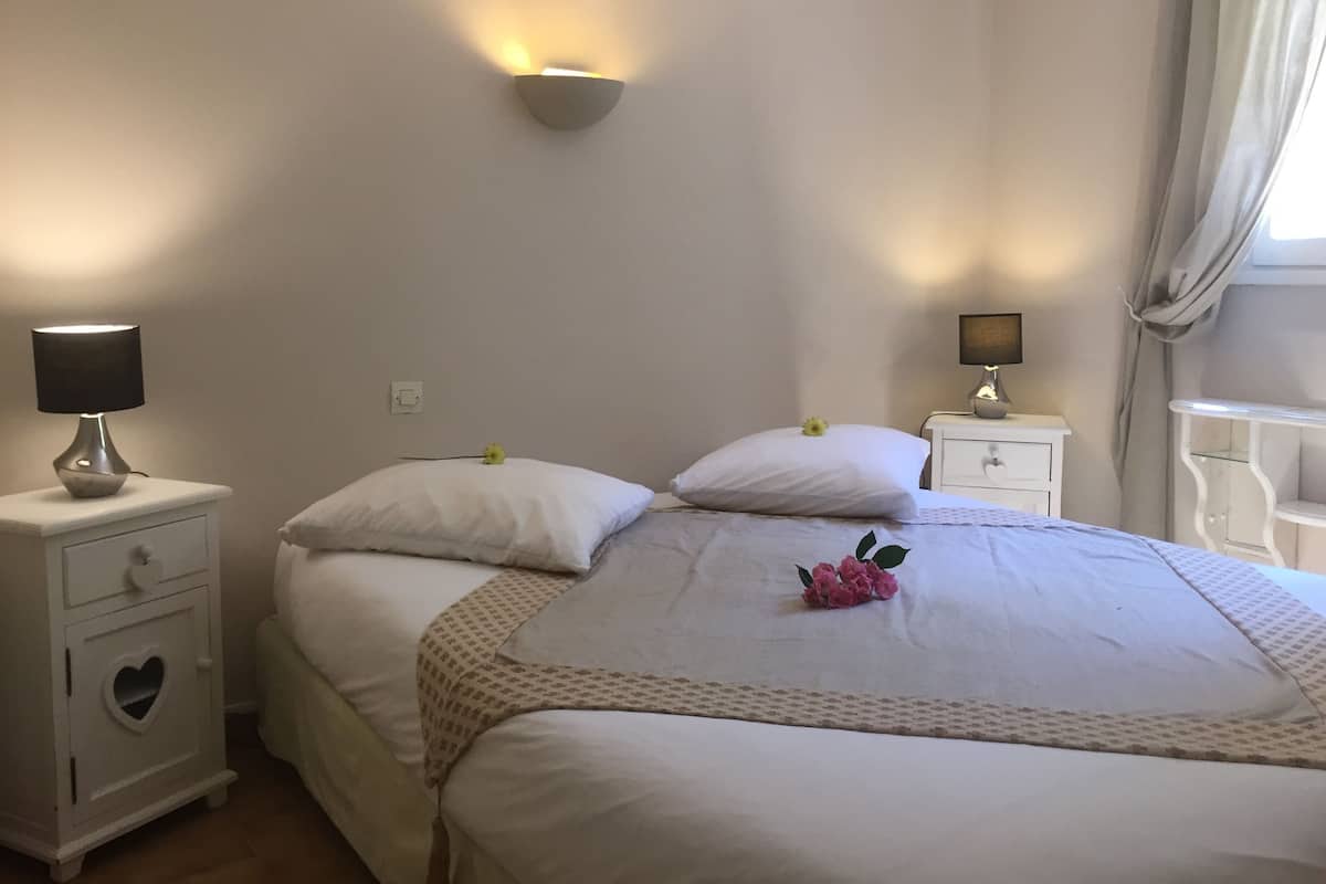 Una cama bien hecha con un hueco en forma de corazón en la cabecera, una lámpara de noche y una lámpara de pared.